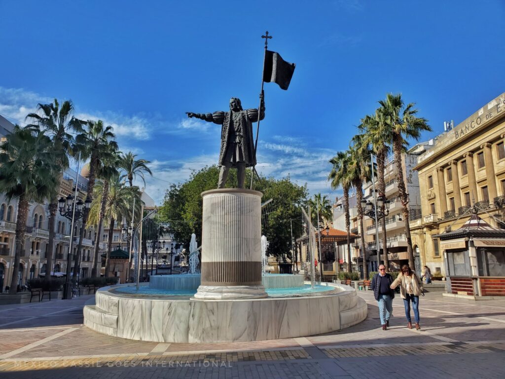 statue of Columbus holding a flag - small ornamental water feature behind him. palm trees lining the plaza