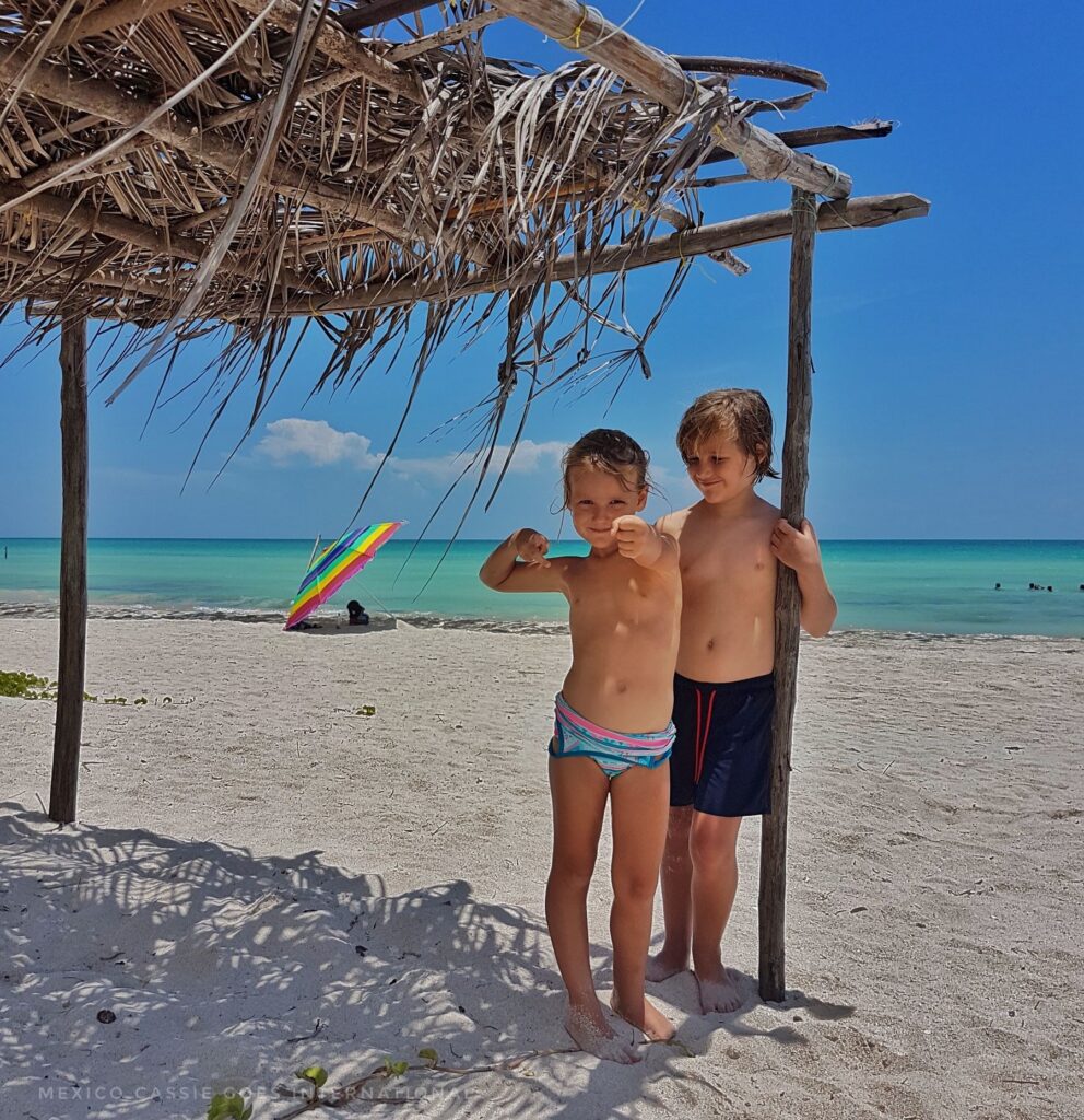 two small children standing under a thatched sun shelter on a beach - white sand, green sea, blue sky, one beach umbrella visible