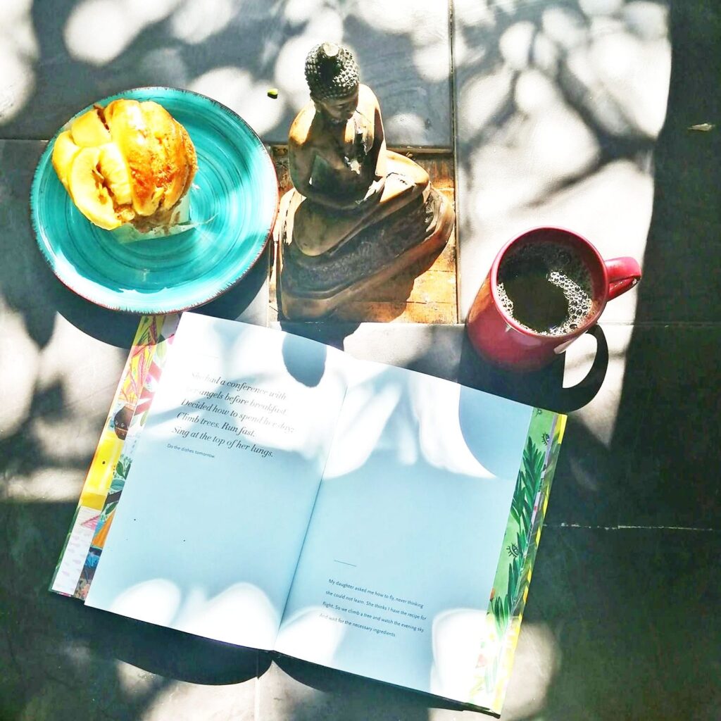 open book on table, red cup of coffee, muffin on a plate and a statue of buddha 