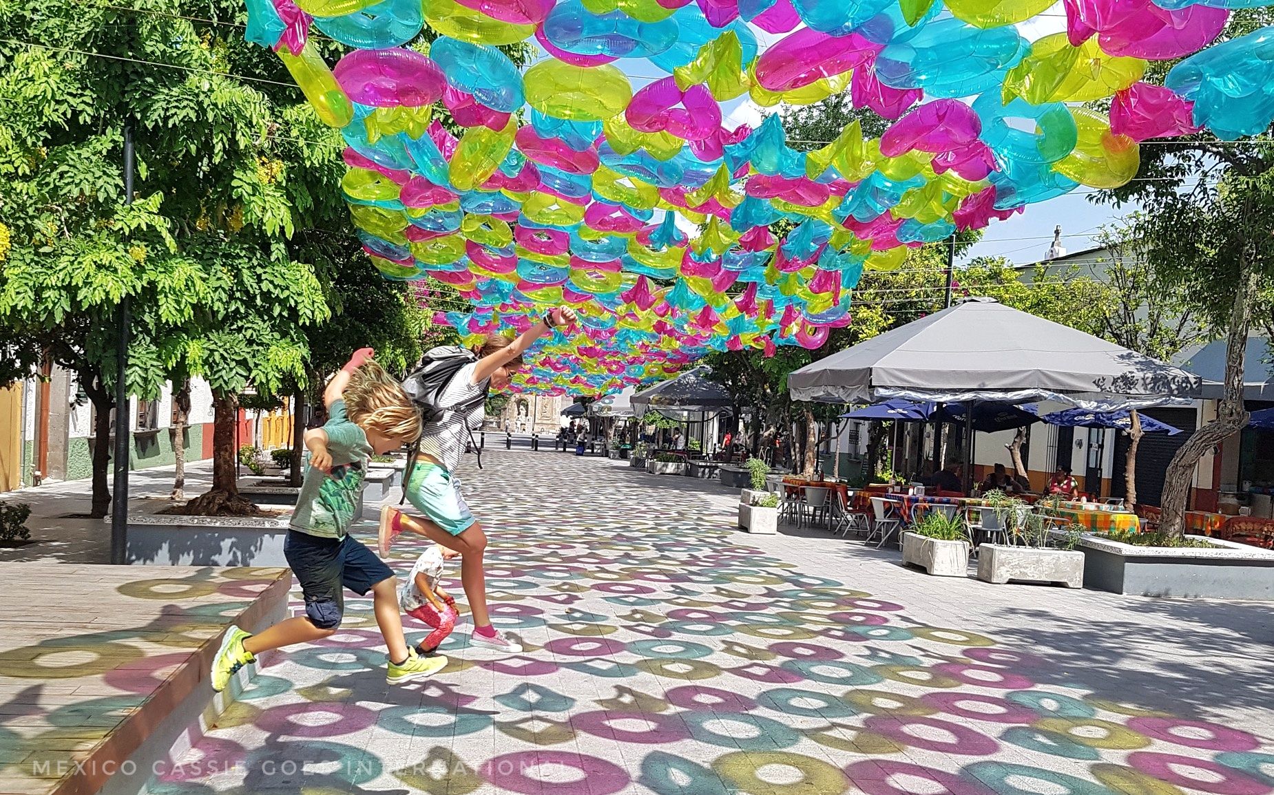 adult and two kids jumping into road - road is covered with inflatable rings (blue, yellow, pink), that cast colourful shadows on the ground
