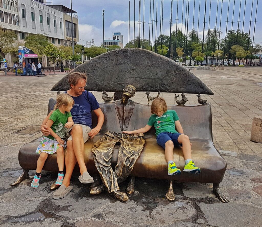adult male and 2 kids sitting on a sculpture bench - flat human between them - part of the sculpture