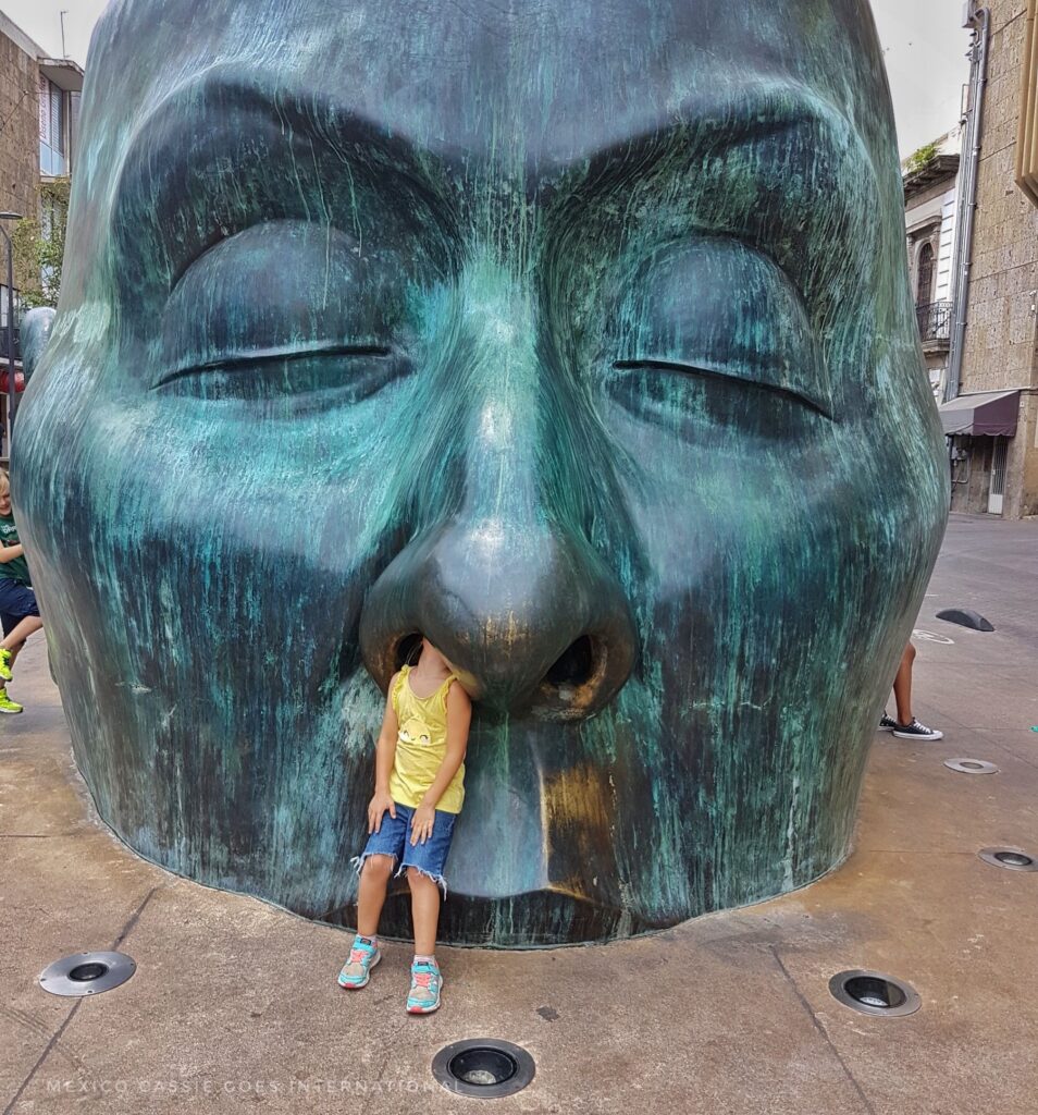 sculpture of a giant head - child in yellow tshirt standing with her had up one nostril