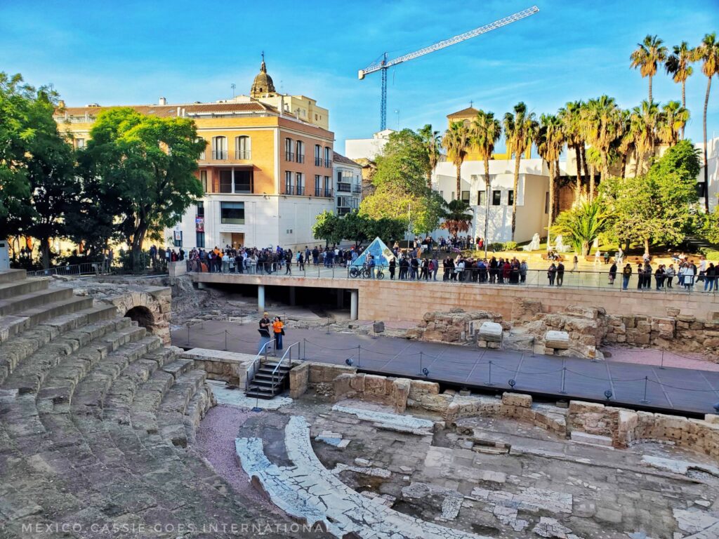 view of the malaga roman theatre - people standing on other side of barrier looking, small glass pyramid and modern buildings behind them
