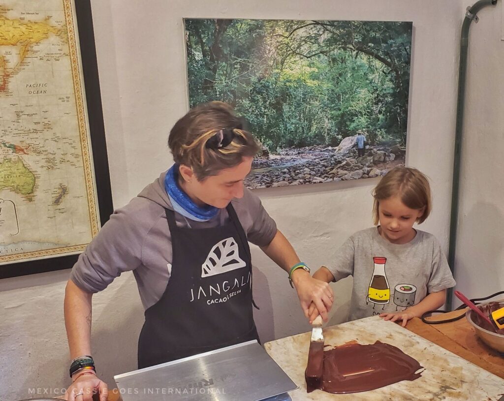 adult in blue apron and child in grey tshirt. Adult is helping child to spread melted chocolate on a marble tray