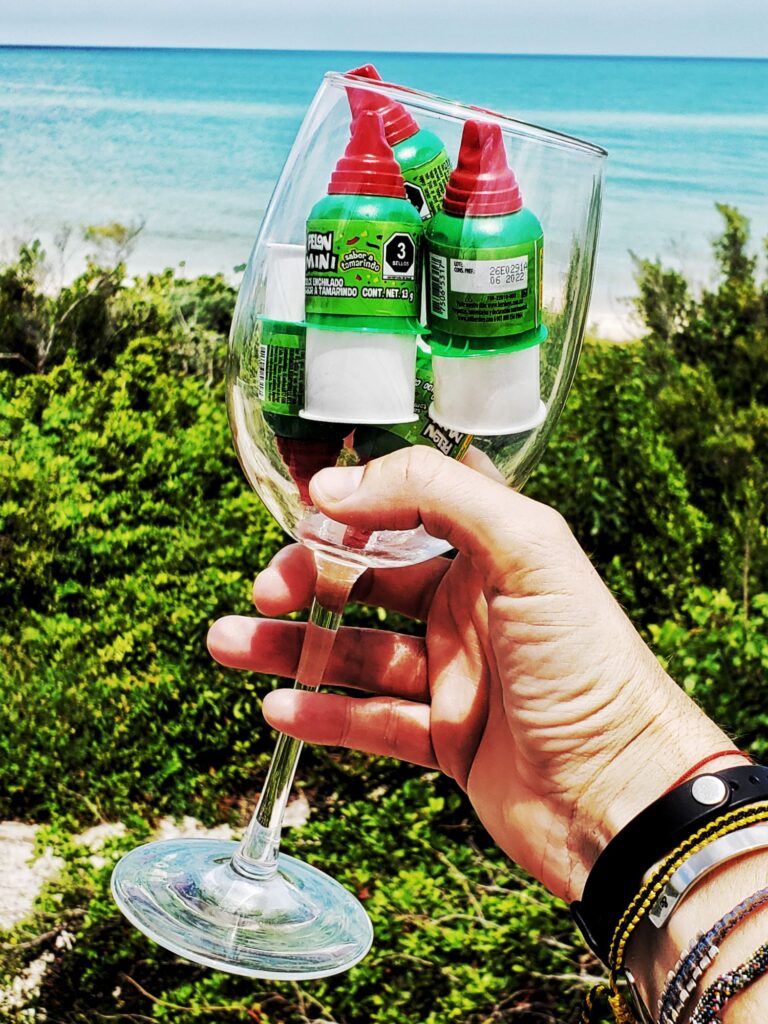 a hand holding a wine glass with 3 pelon pelo rico candy tubes (white, green and red) - beach and beach shrubs visible behind