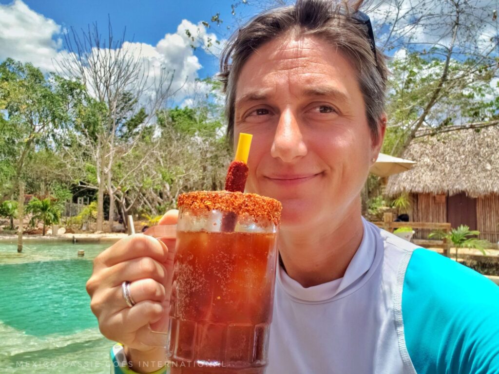 Cassie holding a beer with a tamarind stick in it and spicy salt around the rim - the beer is a michelada so is red