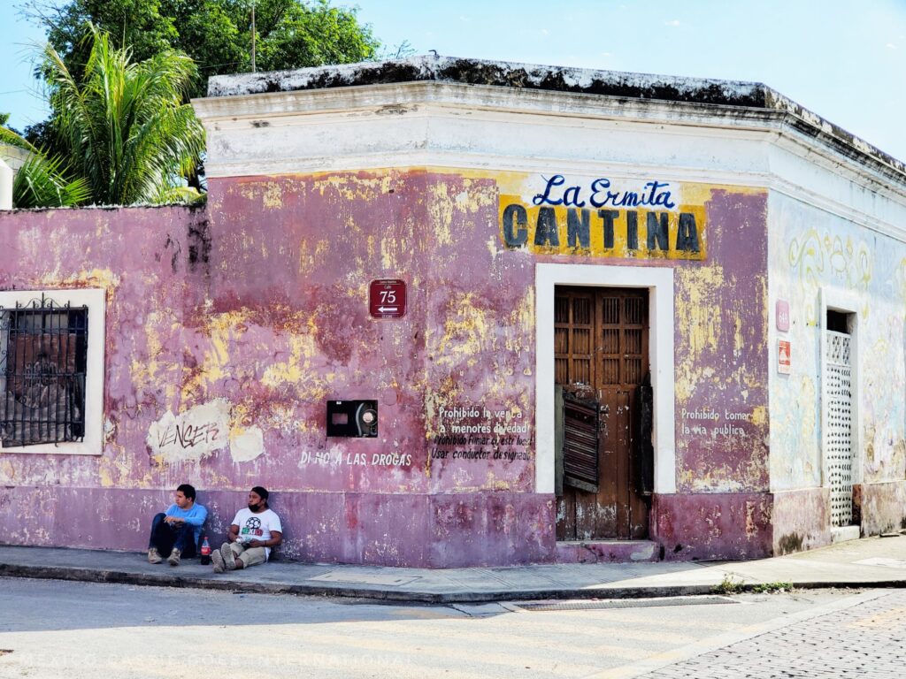 rough pink corner building. 2 men sitting on floor. above door says "La Ermita Cantina"