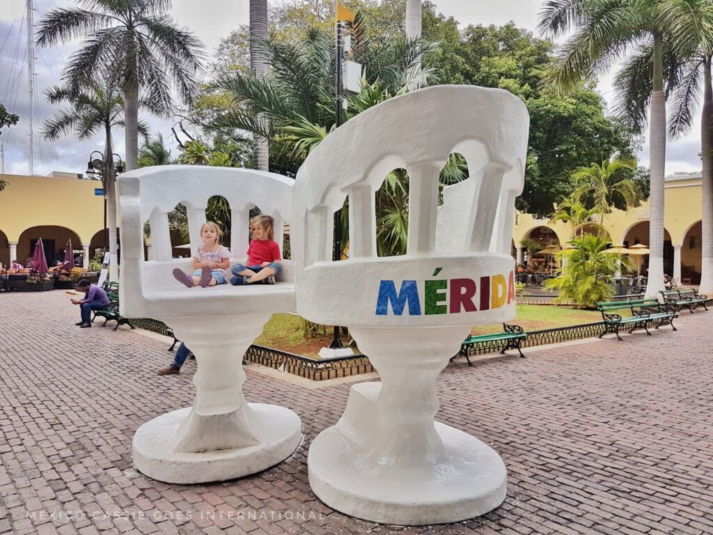 2 giant white chairs on a plaza. one has "Mérida" written on it. 2 small kids sit on the other