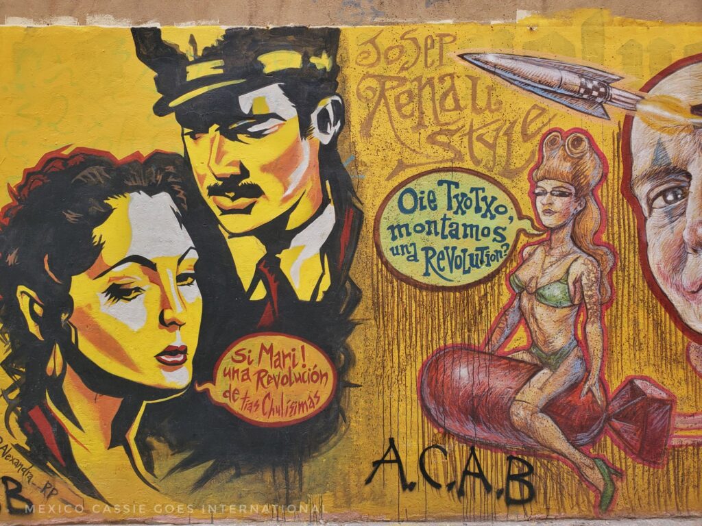 street art. on left, man (chest and head only - in a suit and tie and hat), woman head and neck only. She says, "si Mari! una revolucion de tias chulisimas". On right a woman in a bikini on a bomb saying, "oeie txotxo, montamos una revolution?"