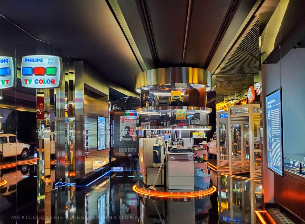 busy modern museum room full of bright lights that are reflected in the floor  - in centre is a circular display with modern appliances such as a vacuum, washing machine and fridge. car reflected on left. telephone box on right and on left also a Philips tv color sign 