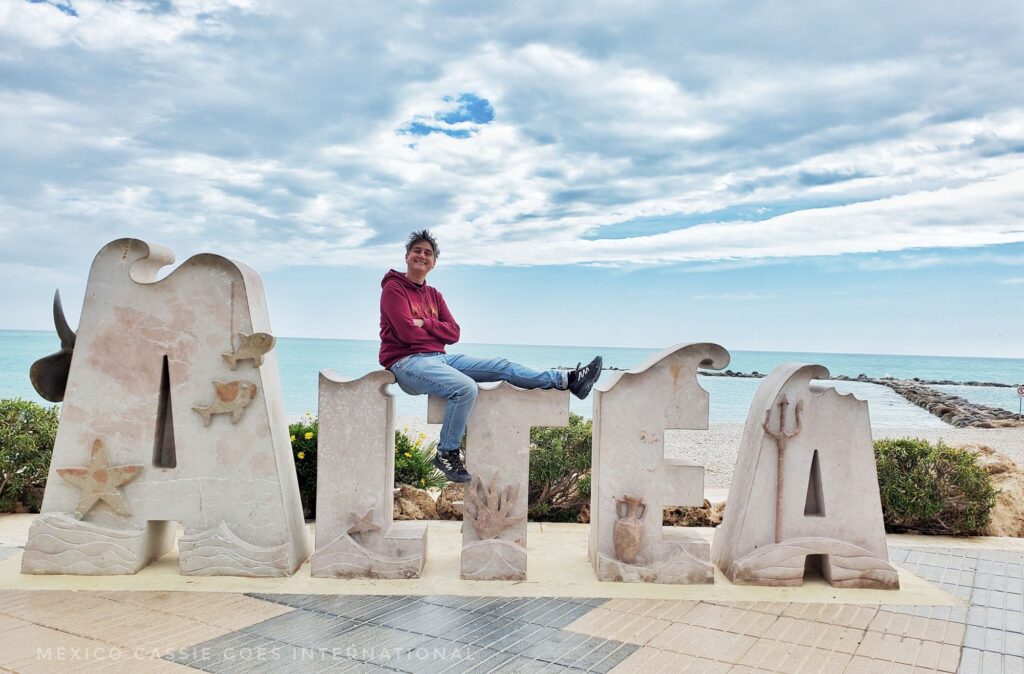 MexicoCassie in a red hoodie sitting on the white oversized letters of Altea in front of the blue ocean