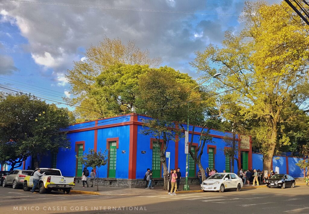 Frida Kahlo's Blue House from the outside - one storey blue building with red trim. trees around, cars on each side of building and people milling around