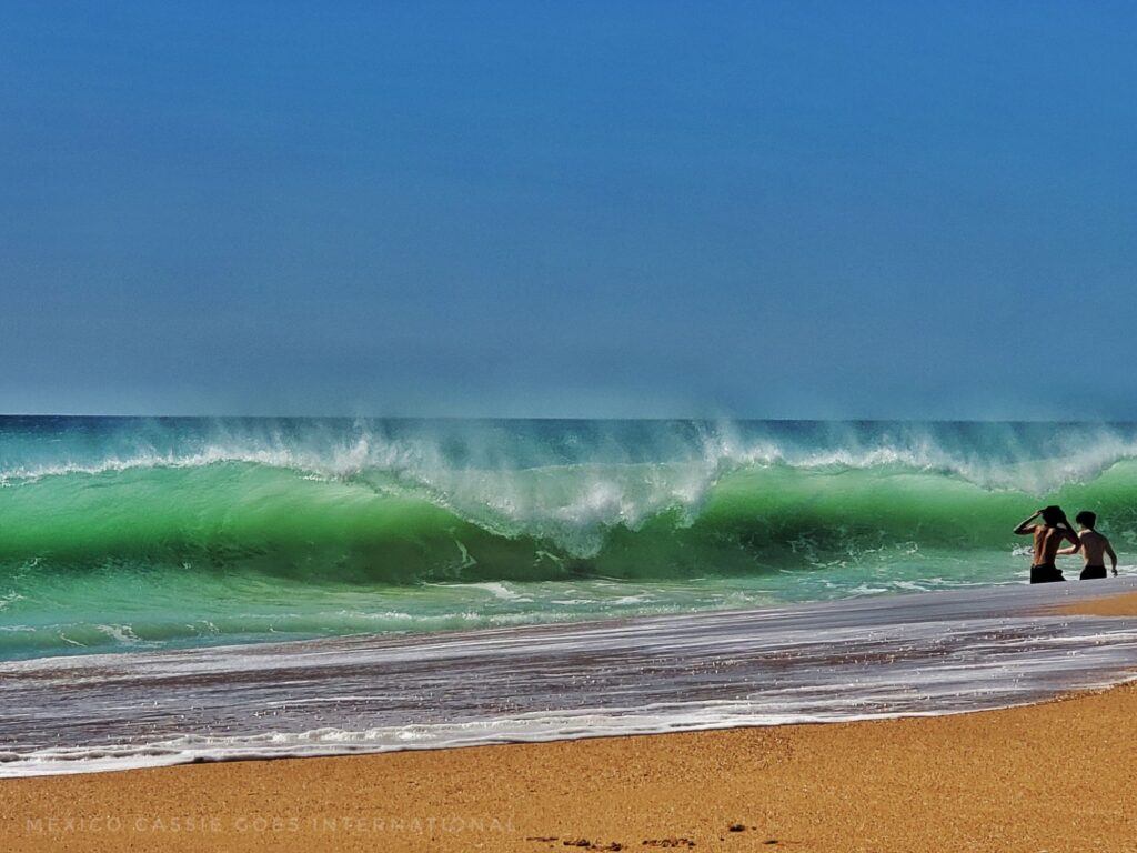green wave crashing on beach, sand and bright blue sky