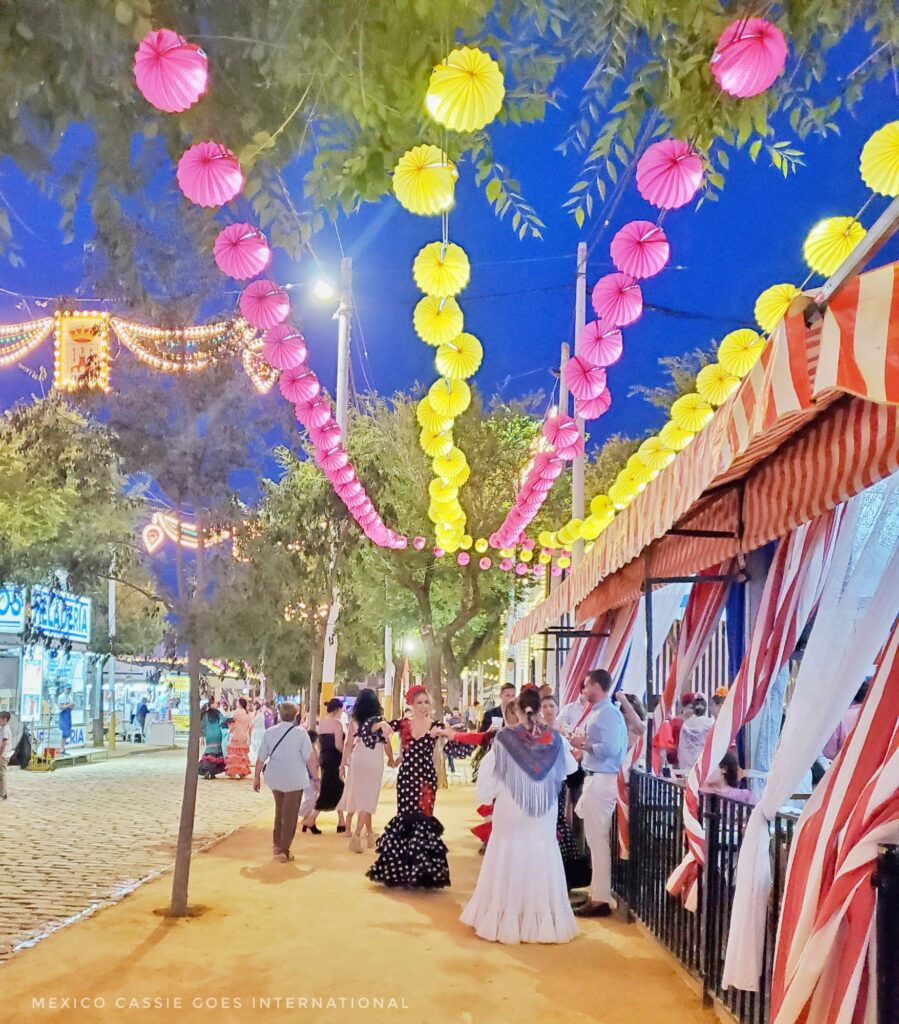 shot of feria scene - people in flamenco dresses, pink and yellow lights strung up and tent open to public