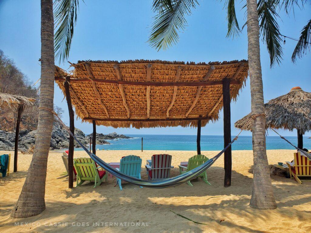 beach scene, brightly coloured wooden chairs under a thatched shade, hammock strung between 2 palm trees