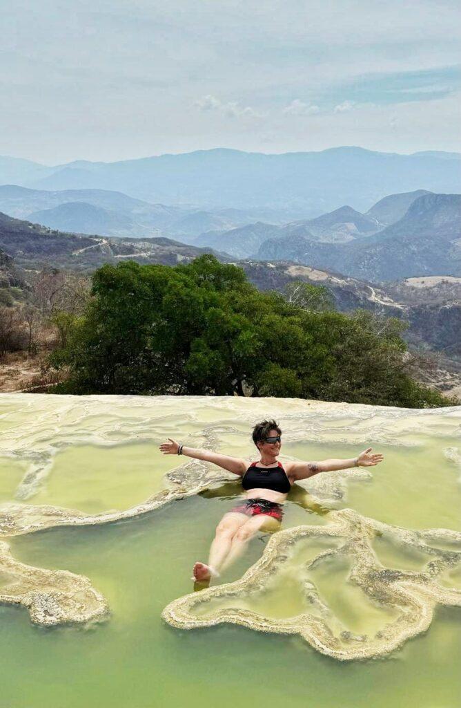 person laying in a small pool of water with hills behind, her arms are outstretched