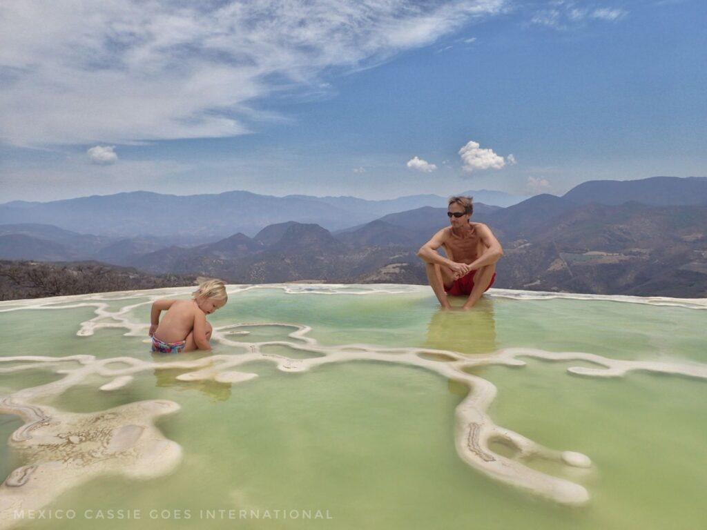 man and small child at hierve el agua- both sitting in the green water, little kids is examining something. gorgeous hills behind