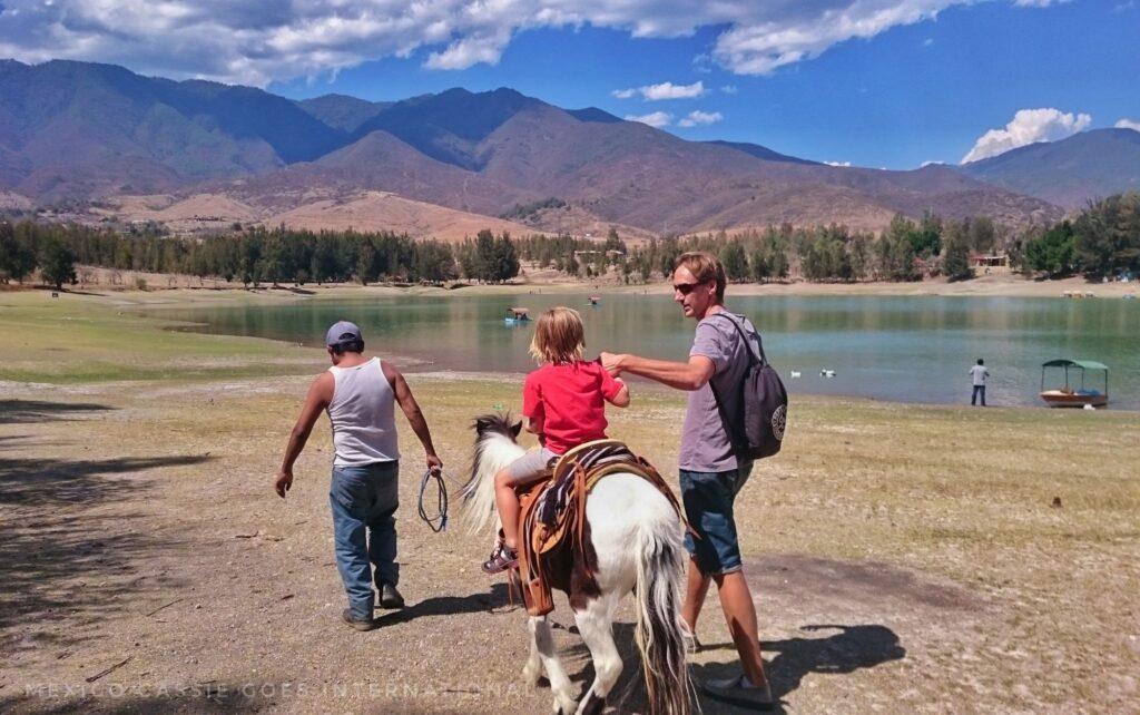 child in red shirt on a small horse being led by 2 men (next to a lake)