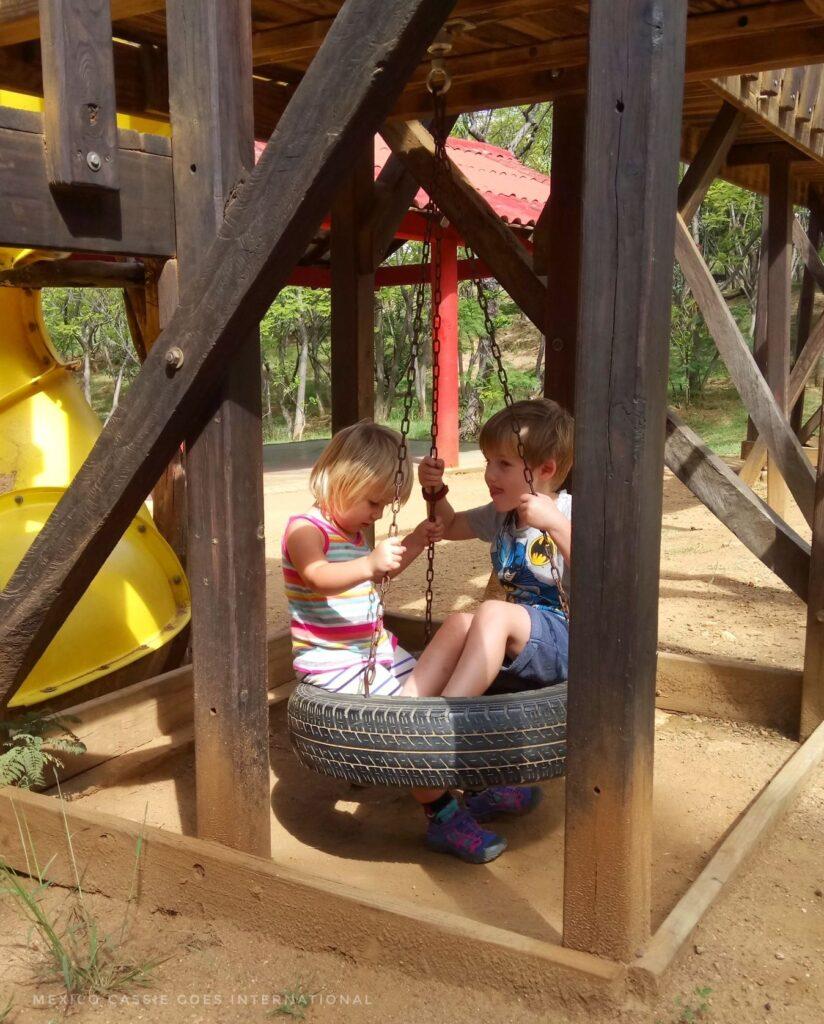 2 small children sitting on a tyre swing under a wooden climbing frame