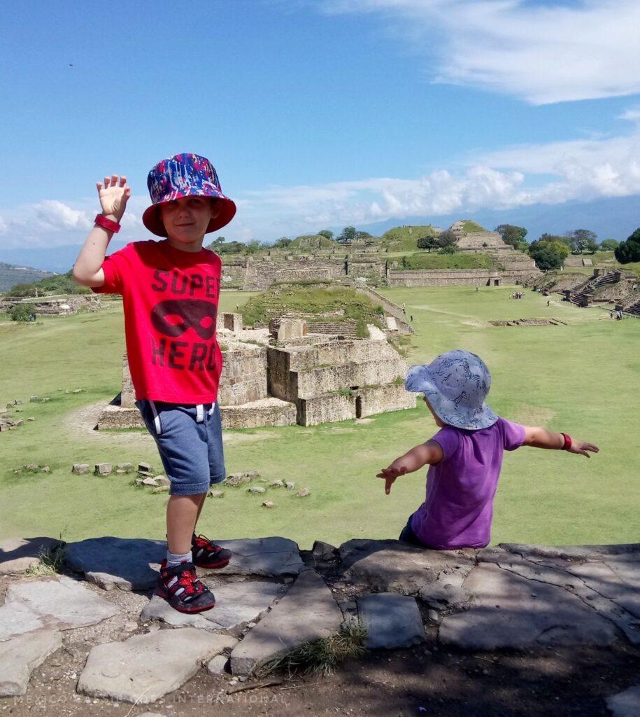 2 small kids (1 in red facing camera, 1 in purple facing away) at top of steps, view of monte alban ruins behind them