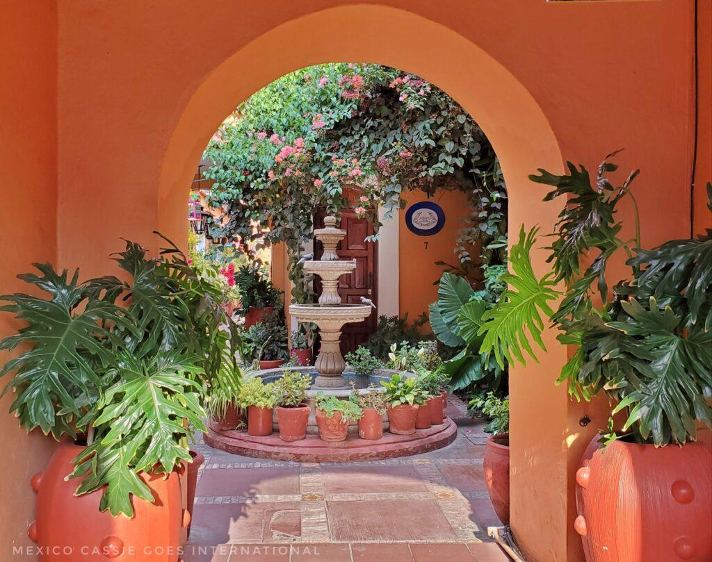archway in orange wall leading onto a fountain, plants all around