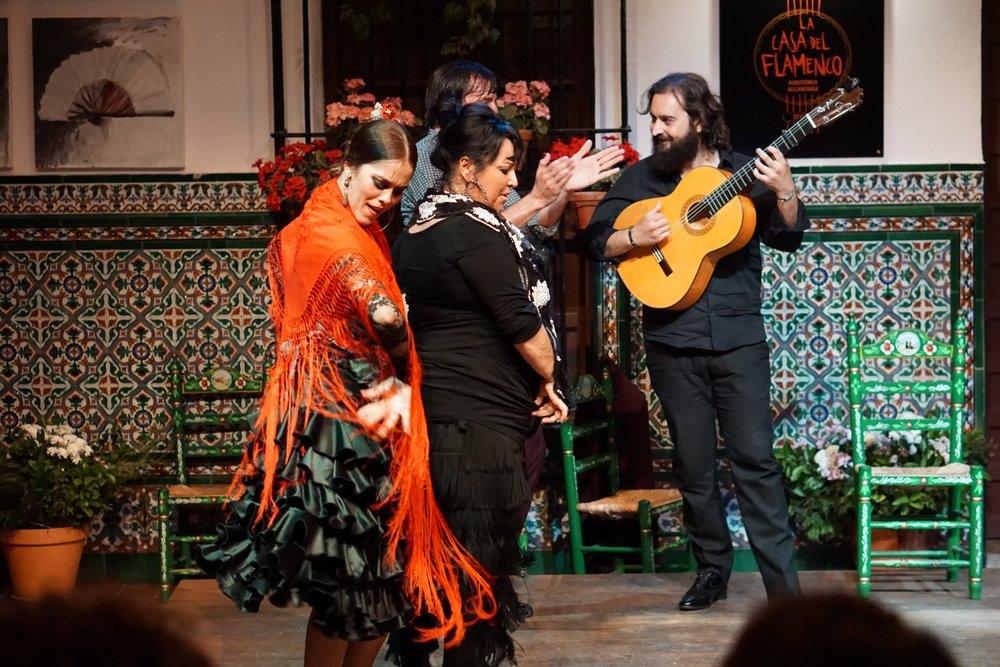 woman in red flamenco dress dancing. man with guitar playing it and another woman in black dancing