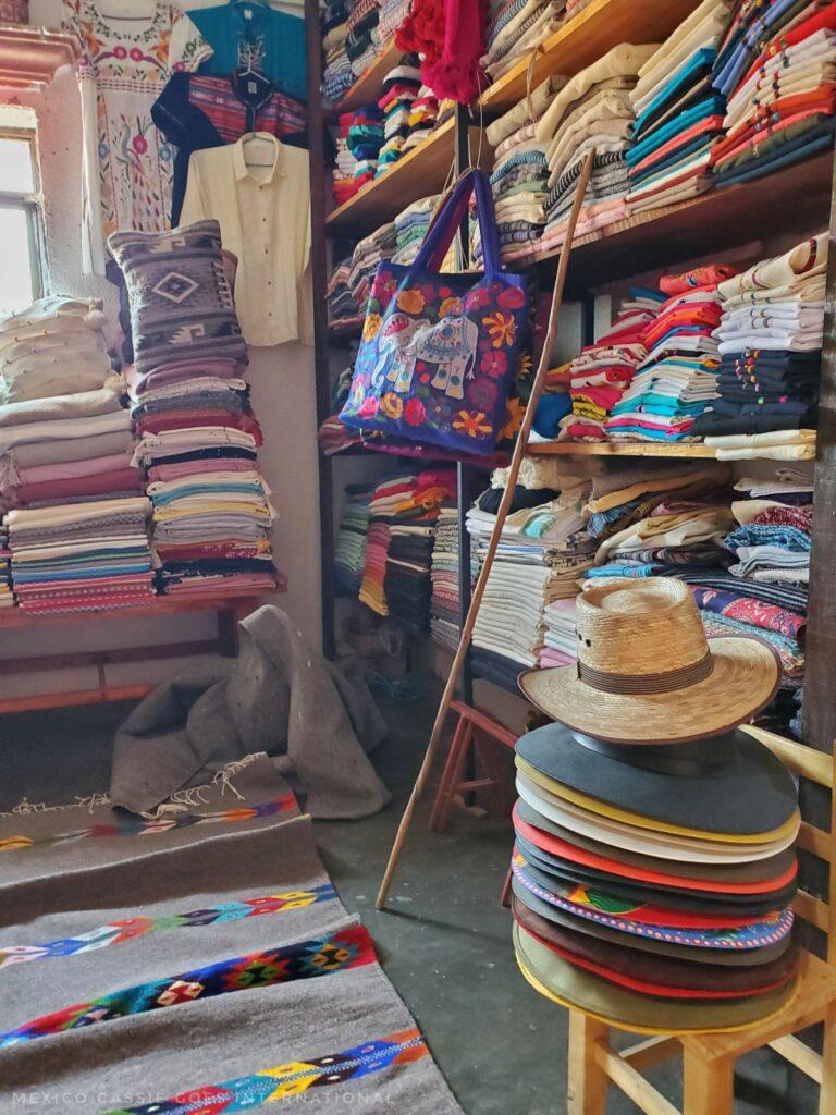 view of inside of shop with piles of rugs and hats. one bag hanging from shelf