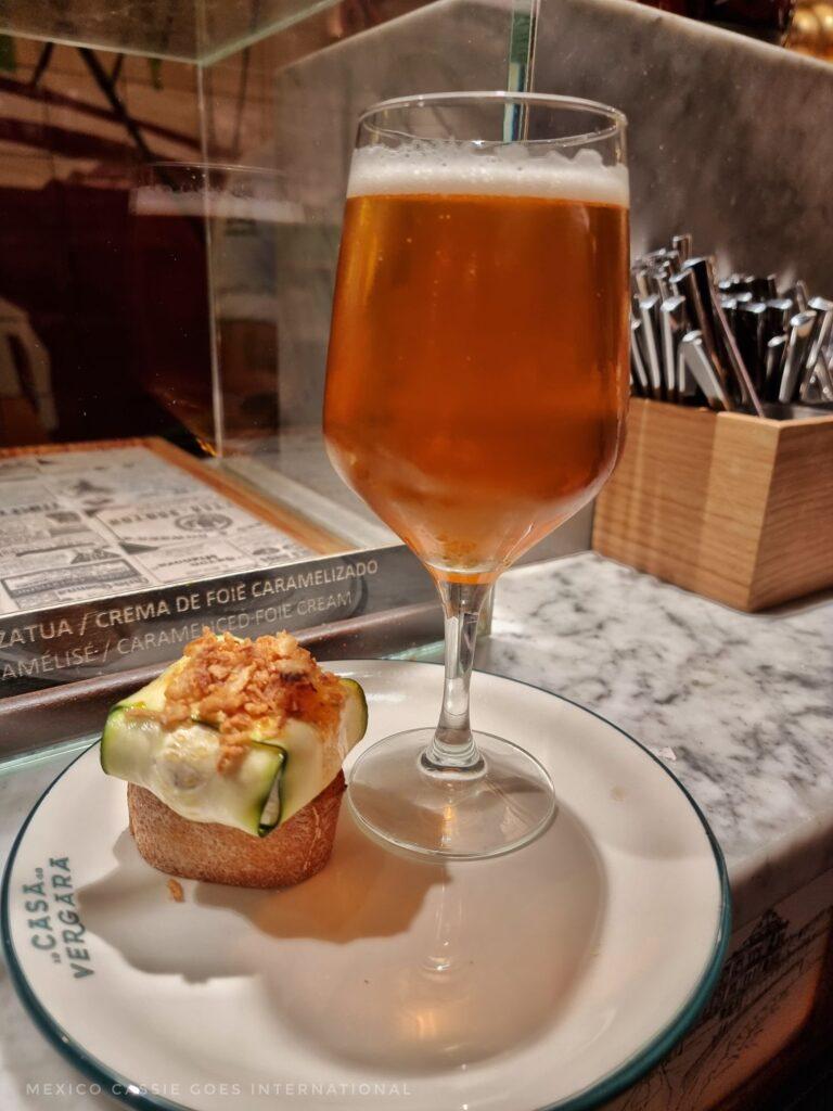 piece of bread with chunk of cheese wrapped in a sliced courgette on a plate next to a beer