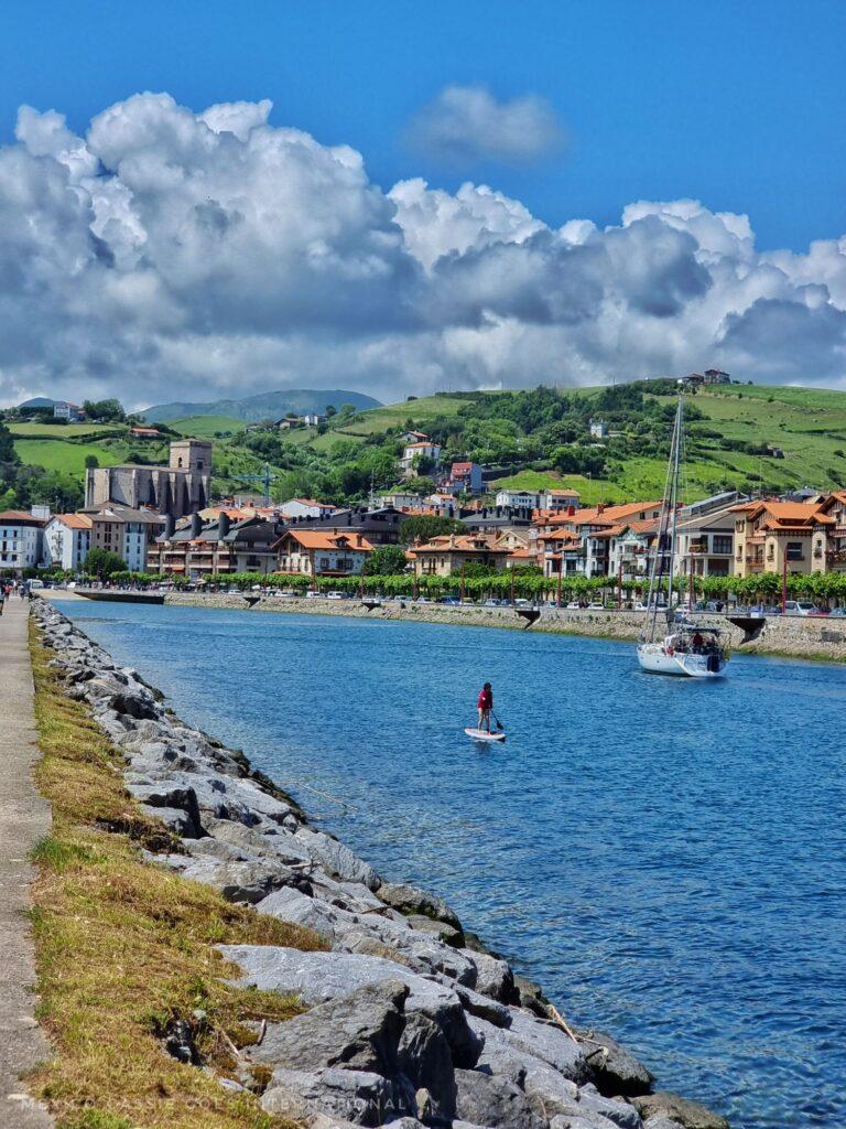 clean looking river with a boat and someone paddle boarding, gorgeous small town behind in hills, big white clouds and blue sky