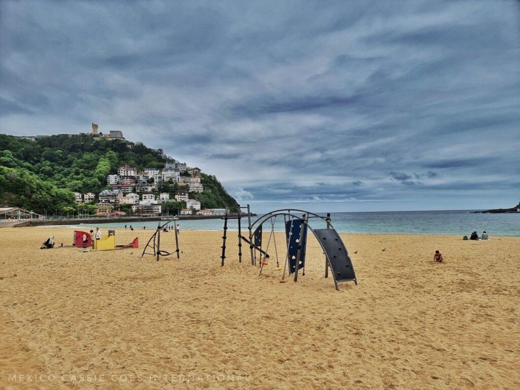 empty sandy beach with some children's play equipment in foreground. green hill in background, moody, cloudy sky