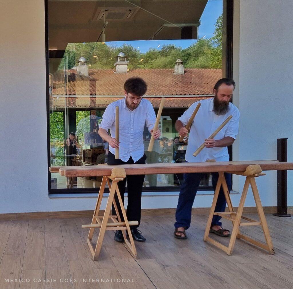 2 men in white shirts playing a large wooden percussion instrument