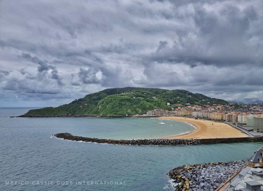 view over playa zurriola  from a nearby viewpojnt - curved beach, breaker, river mouth visible and green hill on far side of beach