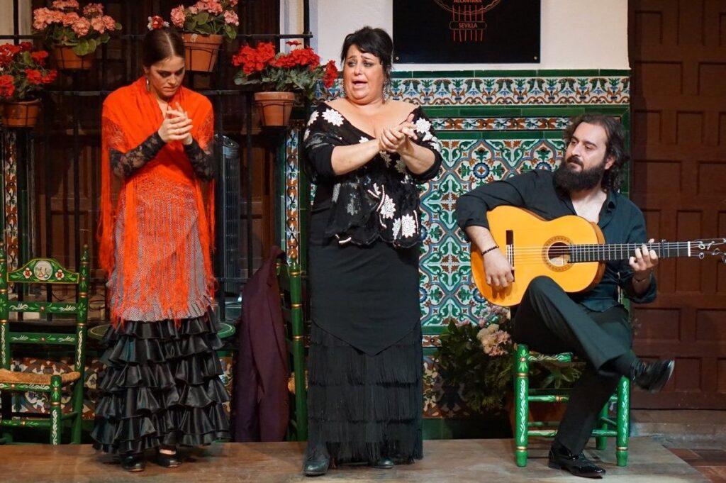 2 women in flamenco dresses clapping hands next to man playing flamenco guitar while seated