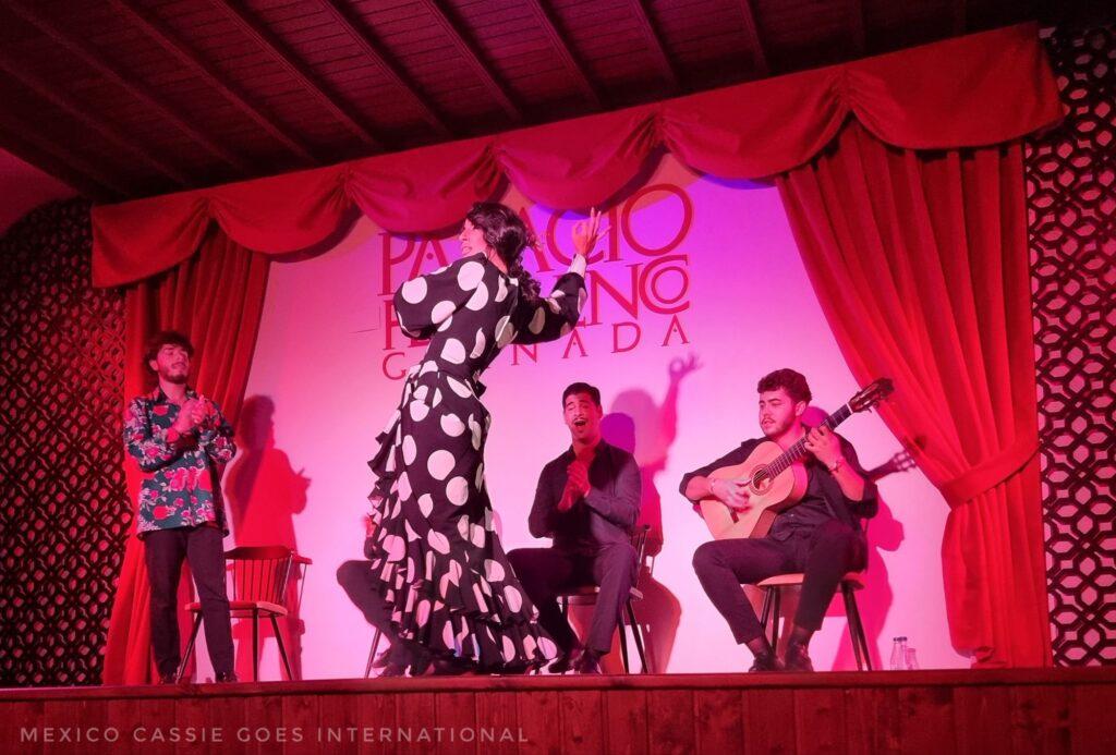 woman dancing on stage in a black and white polka dot dress. Guitarist, singers and male dancer behind her. Photo is bathed in red light