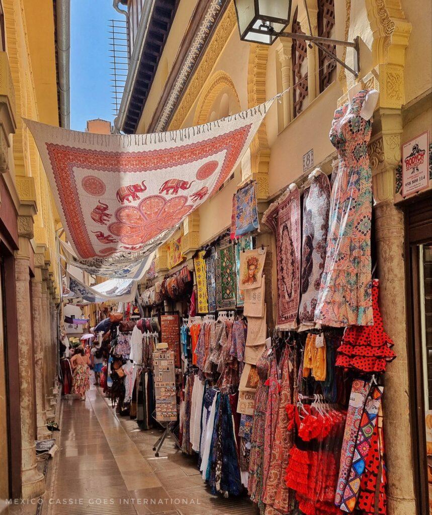 narrow, pedestrianised shopping street with sarongs hung to provide shade, goods displayed outside shops on the walls