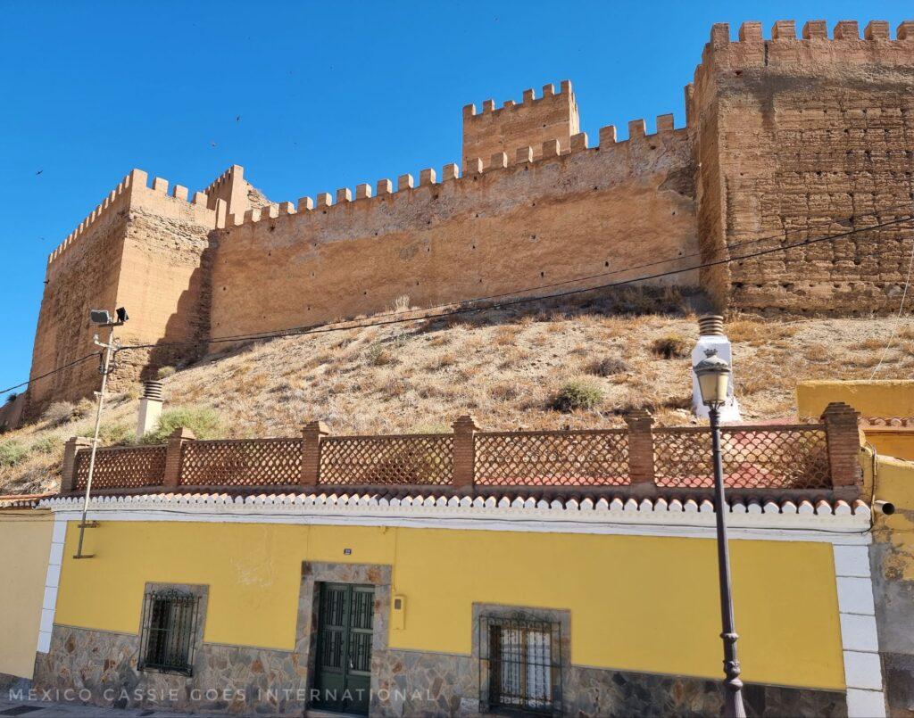 Old fortress wall on hill, small yellow fronted house in hill under fortress