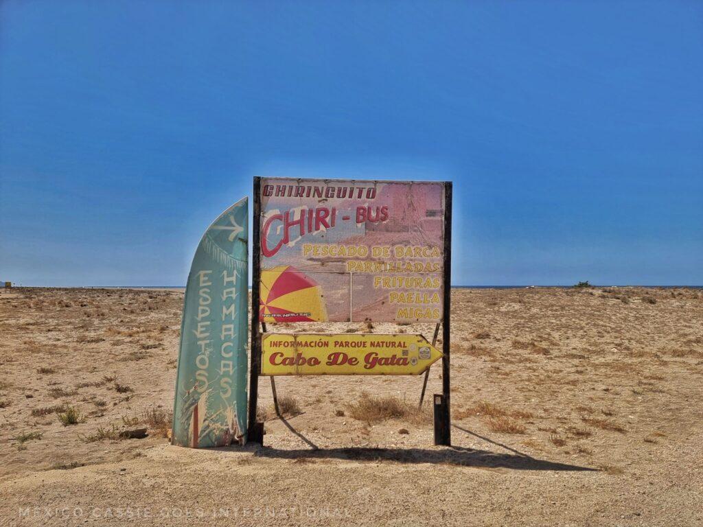 faded sign for a beach bar with a wooden surf board next to it. blue sky and sand