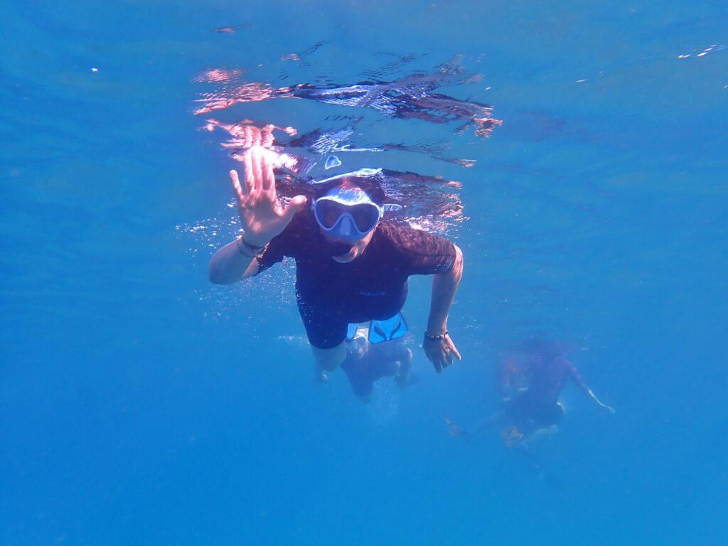 person in wetsuit and snorkel gear (in water, swimming) waving at the camera