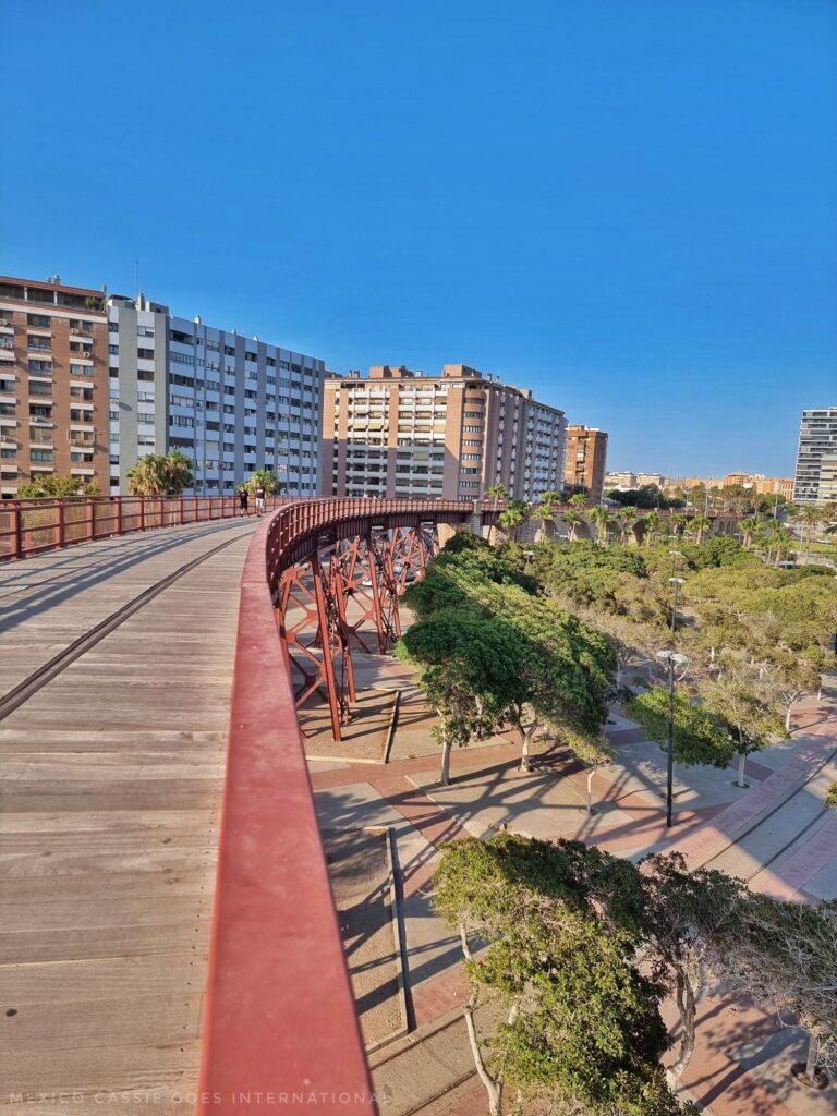 El Cable Inglés, a wooden bridge like structure with red sides, curving into distance, tall buildings on left and park with palm trees on right
