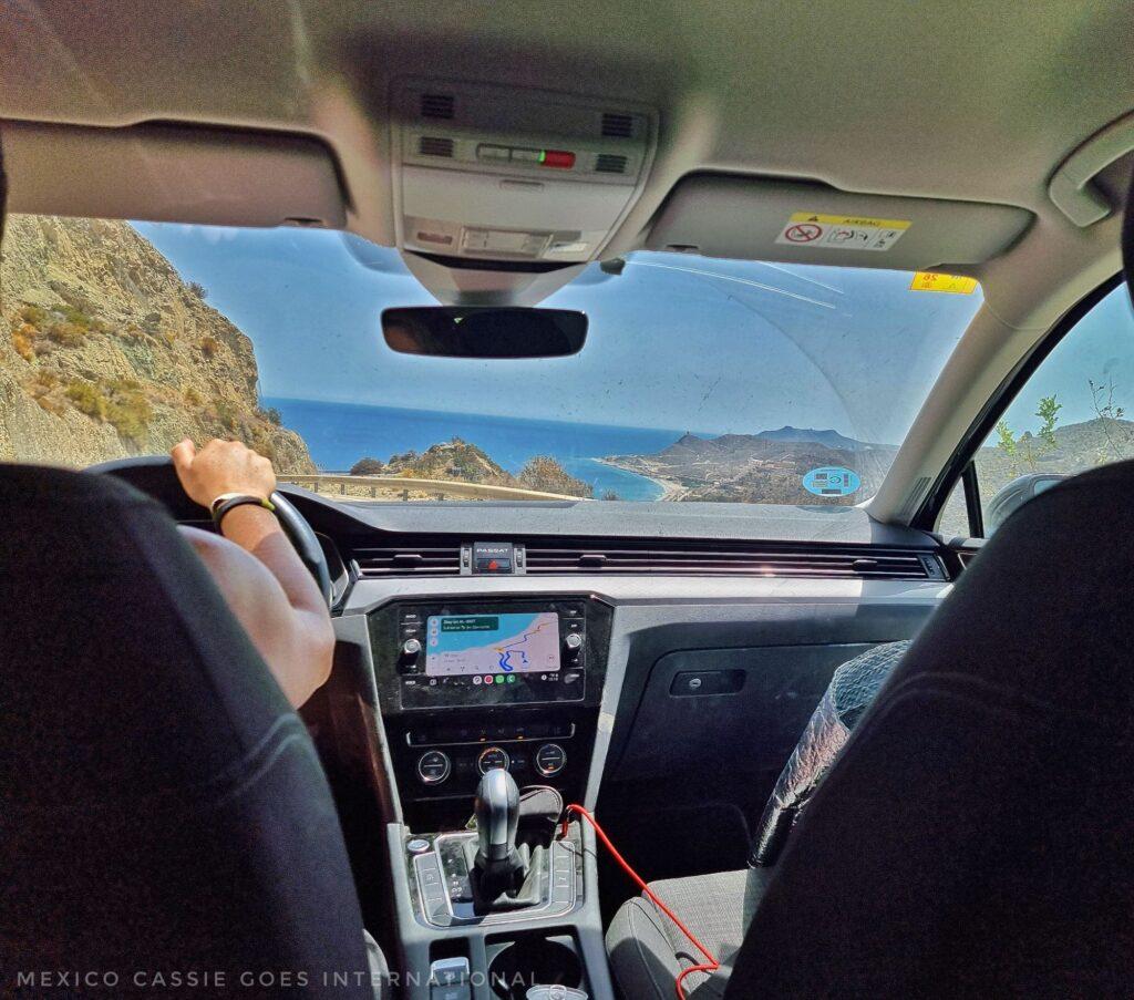 photo taken from back seat of car - see through front window to ocean in front- can also see the right hand and arm of the driver on the left. gps map is also visible although small