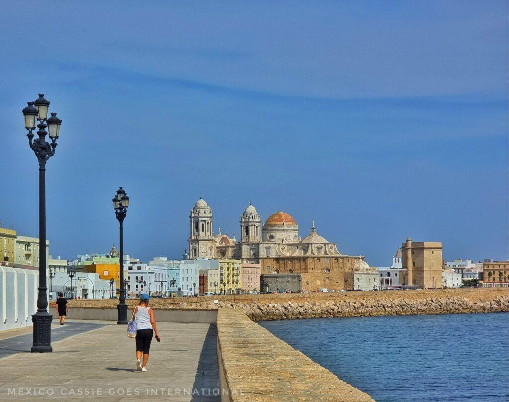 person in distance walking along boardwalk. blue sea to right, cathedral in distance on left