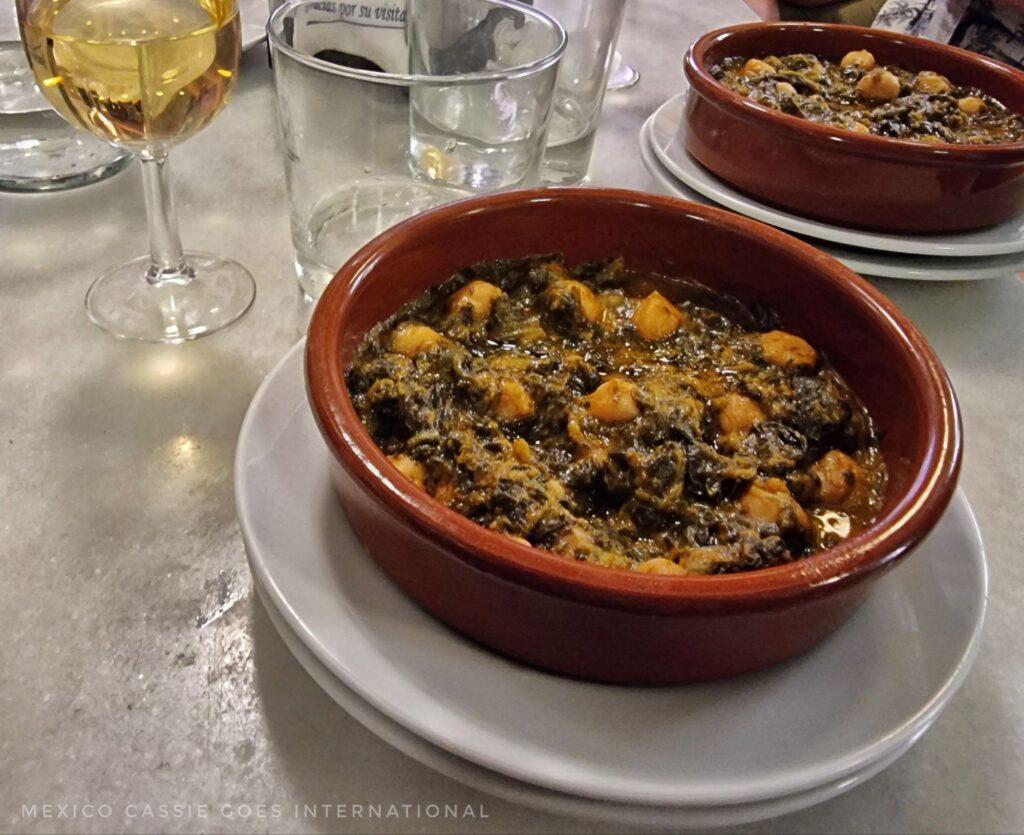 small brown dish with spinach and chickpeas (cooked) in it. Glasses around on table