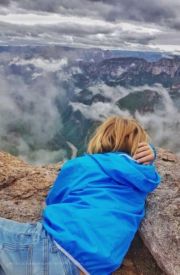 child in jeans and blue jacket lying on rock overlooking a green and cloudy canyon