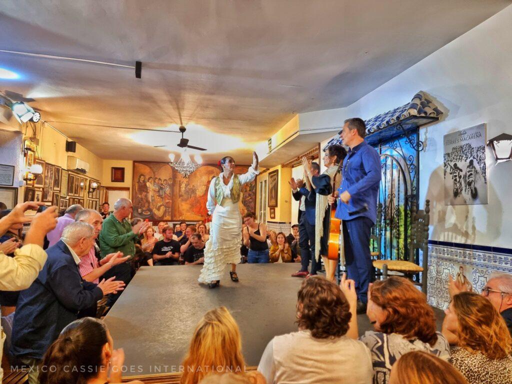 flamenco dancer in white on stage, musicians standing in a line, audience watching