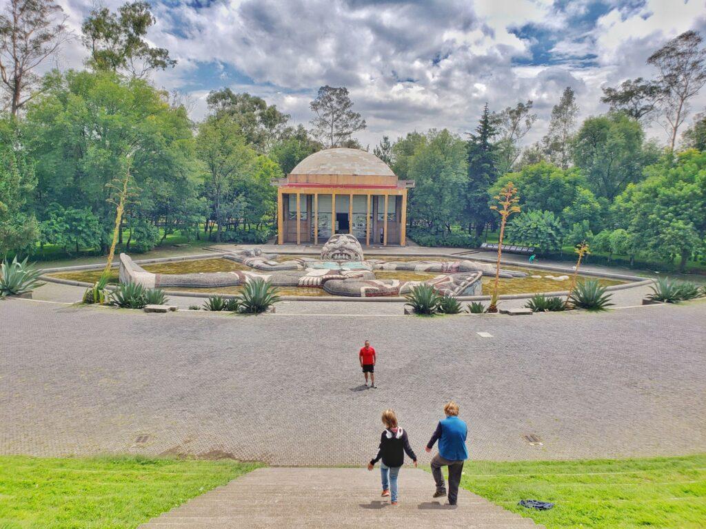 kids running down steps in a park - gold domed building in front of them and before that a pond with a prone sculpture of tlaloc