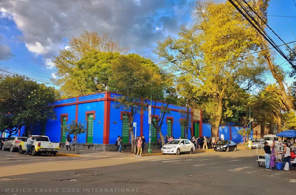 view of outside of frida kahlo's blue house - blue single storey building, trees and people around it