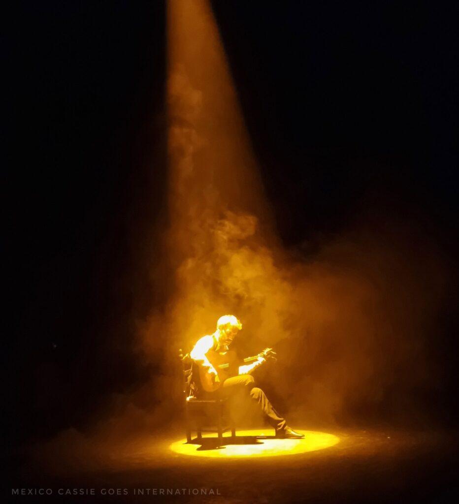 guitarist sitting alone under a yellow spotlight on a dark stage. 