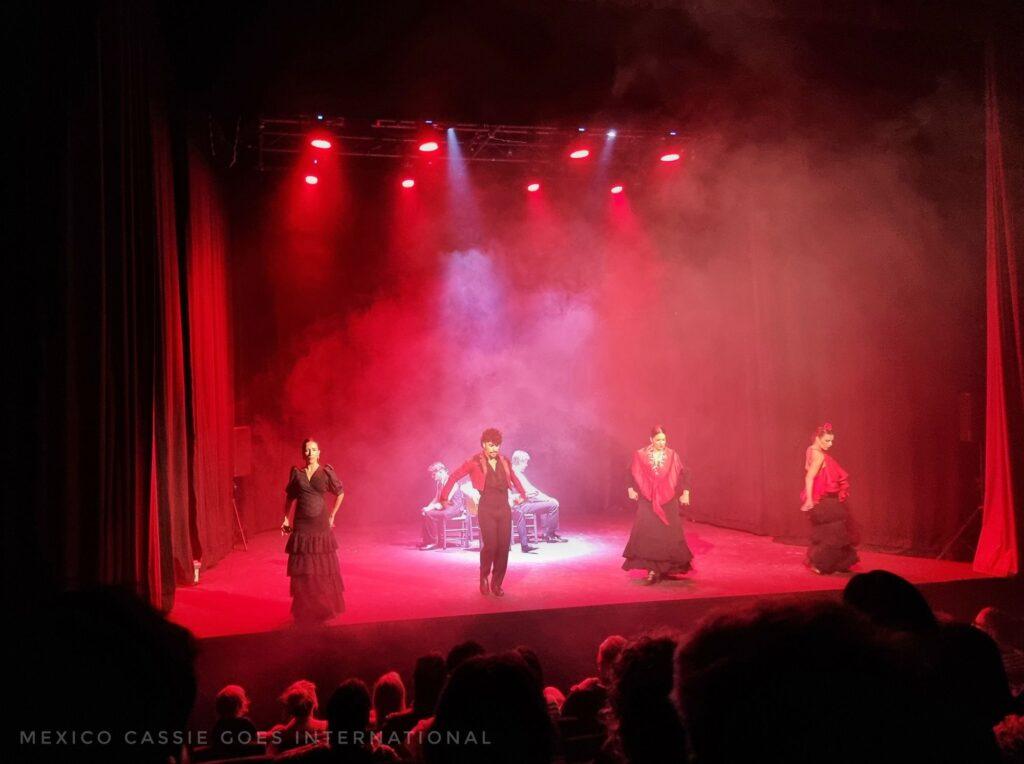 theatre stage bathed in red lights - flamenco dancers (3 women, 1 man)  in a line, 2 singers and guitarist behind, sitting down