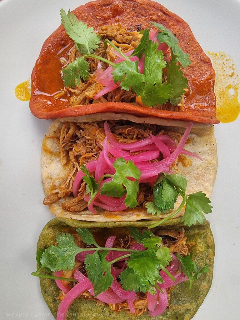 3 tacos on a plate, 1 red, 1 white, 1 green, all with shredded meat, cilantro and red onion