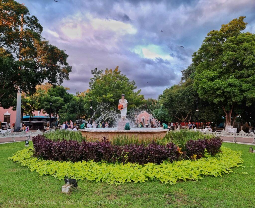 valladolid central plaza - fountain in middle of grass - water spurts towards a statue of a traditionally dressed woman in white huipil holding a jug